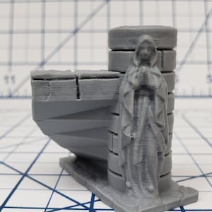 May include: A grey 3D printed figurine of a woman in a robe with her hands clasped in prayer. The figurine is standing on a grey 3D printed base with a pillar behind her.