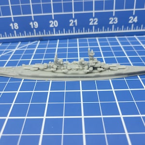 May include: A gray plastic model of a battleship with a detailed design. The model has a long, narrow hull with a raised deck and a tall, slender mast. The model is sitting on a blue gridded surface.