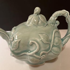May include: A light green ceramic teapot with a floral design. The teapot has a lid and a spout. The handle is shaped like a vine.