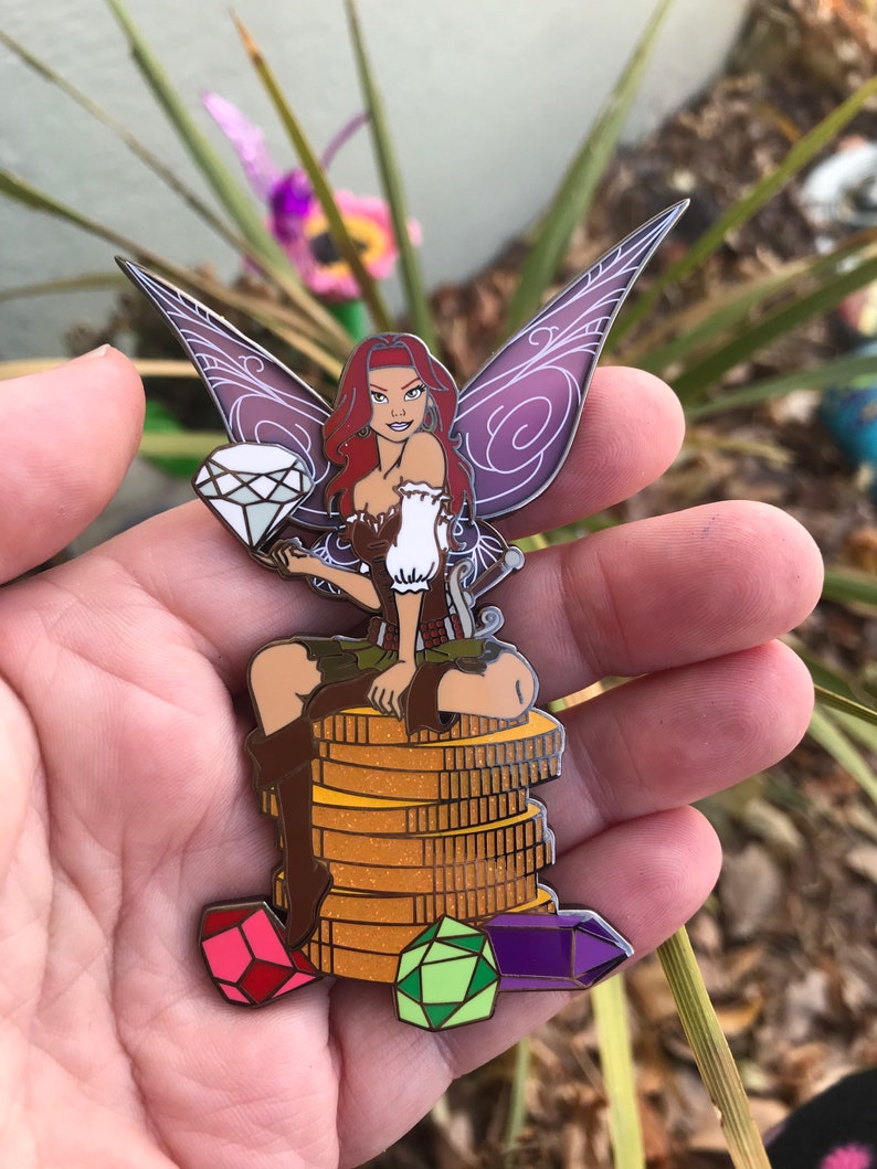 May include: A colorful enamel pin featuring a fairy with red hair, purple wings, and a diamond. The fairy is seated on a stack of gold coins, with colorful gems at the base. The pin is a collectible item.