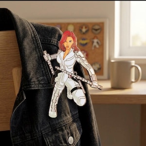 Peut inclure: Épingle en émail Black Widow représentant le personnage en costume blanc et cheveux roux, tenant deux bâtons. L'épingle est fixée à une veste en jean noire.