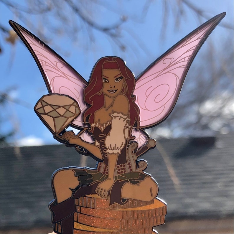 May include: A detailed enamel pin featuring a pirate fairy with red hair, a red headband, and pink wings. She holds a large diamond and sits atop a stack of gold coins. The fairy wears a brown and white outfit with green accents.