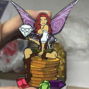 May include: A colorful enamel pin featuring a fairy with red hair, purple wings, and a brown bodice. The fairy is seated on a stack of gold coins and holding a diamond. Several gemstones are at the base.