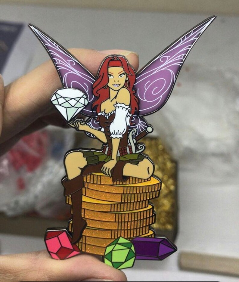 May include: A colorful enamel pin featuring a fairy with red hair, purple wings, and a brown bodice. The fairy is seated on a stack of gold coins and holding a diamond. Several gemstones are at the base.
