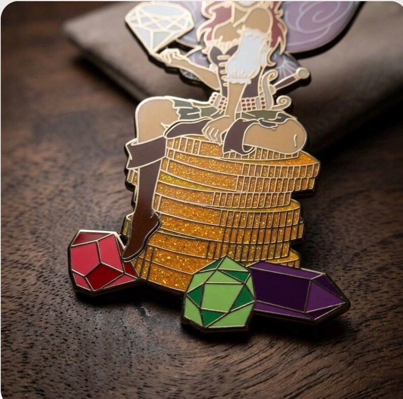 May include: A detailed enamel pin featuring a fairy sitting atop a stack of gold coins. The fairy holds a diamond and is surrounded by red, green, and purple gemstone pins. The pin has a glittery gold background and is set against a dark wood surface.