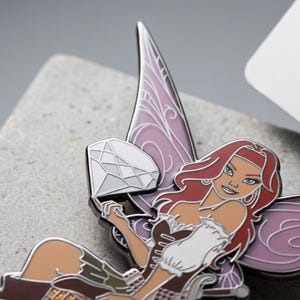 May include: A detailed enamel pin featuring a fairy with red hair, a brown and white dress, and purple wings. The fairy is holding a diamond. The pin has a silver outline and is set against a gray background.