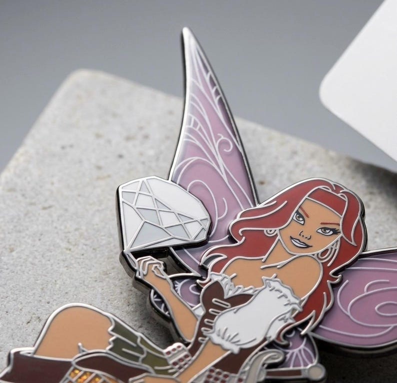 May include: A detailed enamel pin featuring a fairy with red hair, a brown and white dress, and purple wings. The fairy is holding a diamond. The pin has a silver outline and is set against a gray background.
