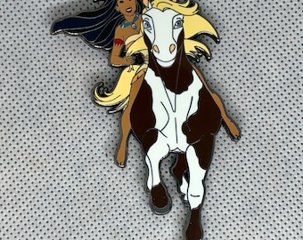 Pode incluir: Um broche com uma imagem de desenho animado de Pocahontas montando seu cavalo, um cavalo pinto marrom e branco com uma juba loira. O cavalo está correndo e Pocahontas está segurando as rédeas.
