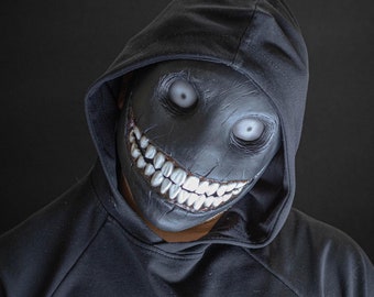 May include: A person wearing a black hooded sweatshirt with a black mask that has a wide, toothy grin and white eyes.