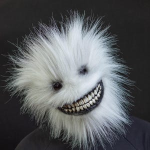 May include: A white, fluffy mask with a wide, unsettling grin and dark eye sockets. The mask is worn over a black top, creating a stark contrast against the black background. The mask's texture appears soft and fluffy.