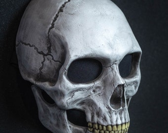 May include: A white skull mask with black eye holes and yellow teeth. The mask is made of a hard plastic and has a realistic look.