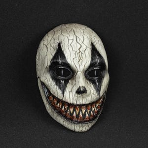 May include: A full-face mask featuring a cracked, off-white surface texture. The design includes sharp black diamond-shaped markings around the eye sockets and a wide, toothy grin with dark, blood-stained detailing along the gum line and teeth.