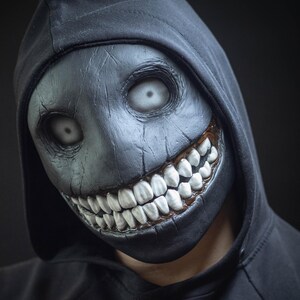 May include: A person wearing a black hooded sweatshirt with a creepy, black and white mask. The mask has a wide, toothy grin and large, white eyes.