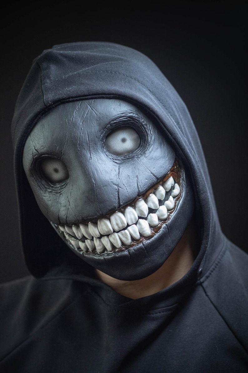 May include: A person wearing a black hooded sweatshirt with a creepy, black and white mask. The mask has a wide, toothy grin and large, white eyes.
