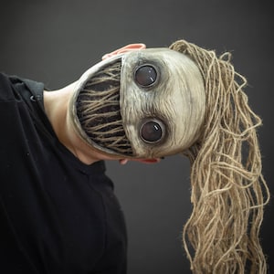 Puede incluir: Una persona lleva una máscara beige espeluznante con una amplia sonrisa de dientes y ojos grandes y oscuros. La máscara tiene una larga peluca marrón y fibrosa.