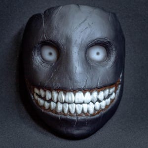May include: A black and white creepy mask with a wide, toothy grin and white eyes. The mask is made of a textured material and has a realistic look.