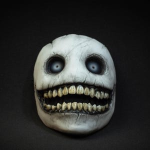 May include: A white, cracked mask with large, hollow eyes and a wide, toothy grin. The mask has a weathered appearance, with visible cracks and shadows, creating a spooky aesthetic. The mask is set against a black background.