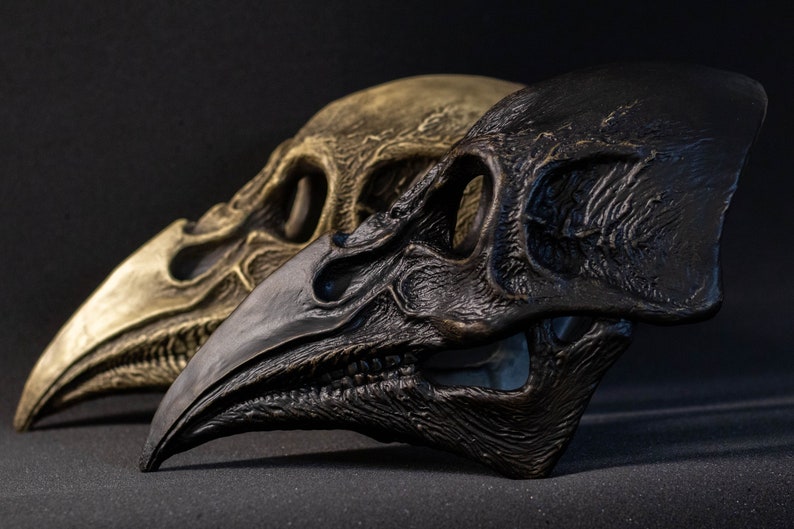 May include: Two bird skull sculptures. One is a metallic gold color, and the other is a dark, matte black. Both have detailed textures and are set against a dark background. The skulls are side by side, showcasing their intricate designs.