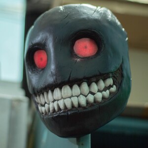 May include: A black mask with a wide, toothy grin and glowing red eyes. The mask is made of a textured material and has a realistic appearance.