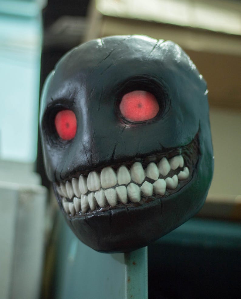 May include: A black mask with a wide, toothy grin and glowing red eyes. The mask is made of a textured material and has a realistic appearance.