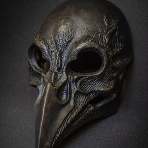 May include: A dark, weathered mask with a bird-like beak and skull-like features. The mask is primarily black with bronze highlights, and has large eye openings. The mask is likely for costume or decorative purposes.