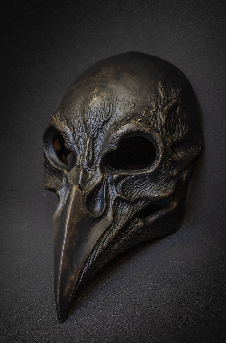 May include: A dark, weathered mask with a bird-like beak and skull-like features. The mask is primarily black with bronze highlights, and has large eye openings. The mask is likely for costume or decorative purposes.