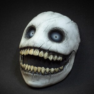 May include: A white mask with a cracked, weathered appearance. The mask features large, hollow eyes with blue irises and a wide, toothy grin. The overall design evokes a sense of the macabre and is suitable for cosplay or costume use.