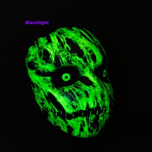 May include: A full-face mask featuring a vibrant neon green marbled pattern against a deep black background. The mask displays large, hollow eye sockets with glowing green circular pupils and a jagged, stitched mouth design. The text Blacklight is visible.