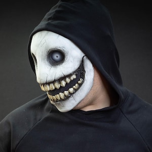 May include: A white and black mask with a sinister grin and large teeth. The mask has a cracked texture and is worn with a black hooded sweatshirt. The mask's eyes are a pale, unsettling color.
