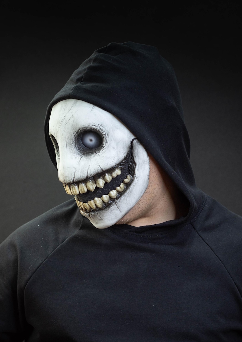 May include: A white and black mask with a sinister grin and large teeth. The mask has a cracked texture and is worn with a black hooded sweatshirt. The mask's eyes are a pale, unsettling color.
