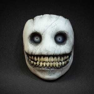 May include: A white mask with a cracked, aged appearance. The mask features large, hollow eyes and a wide, toothy grin. The teeth are yellowed, and the overall design has a slightly unsettling, yet artistic, aesthetic. The mask is likely for cosplay or costume use.