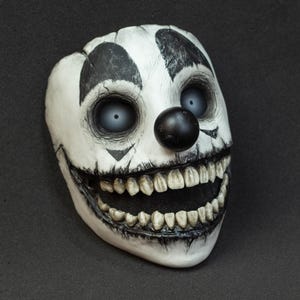 Pode incluir: Uma máscara de palhaço branca com detalhes em preto e um sorriso sinistro. A máscara apresenta olhos grandes e vazios, um nariz preto e marcas pretas ao redor dos olhos e da boca. Os dentes são visíveis e a máscara tem uma aparência rachada e envelhecida.