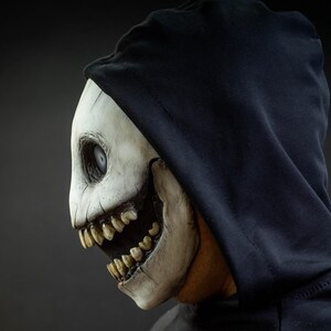 May include: A side profile of a white skull mask with a black hood. The mask has a wide open mouth with sharp teeth and a single, large eye. The mask is worn over a black hooded sweatshirt.