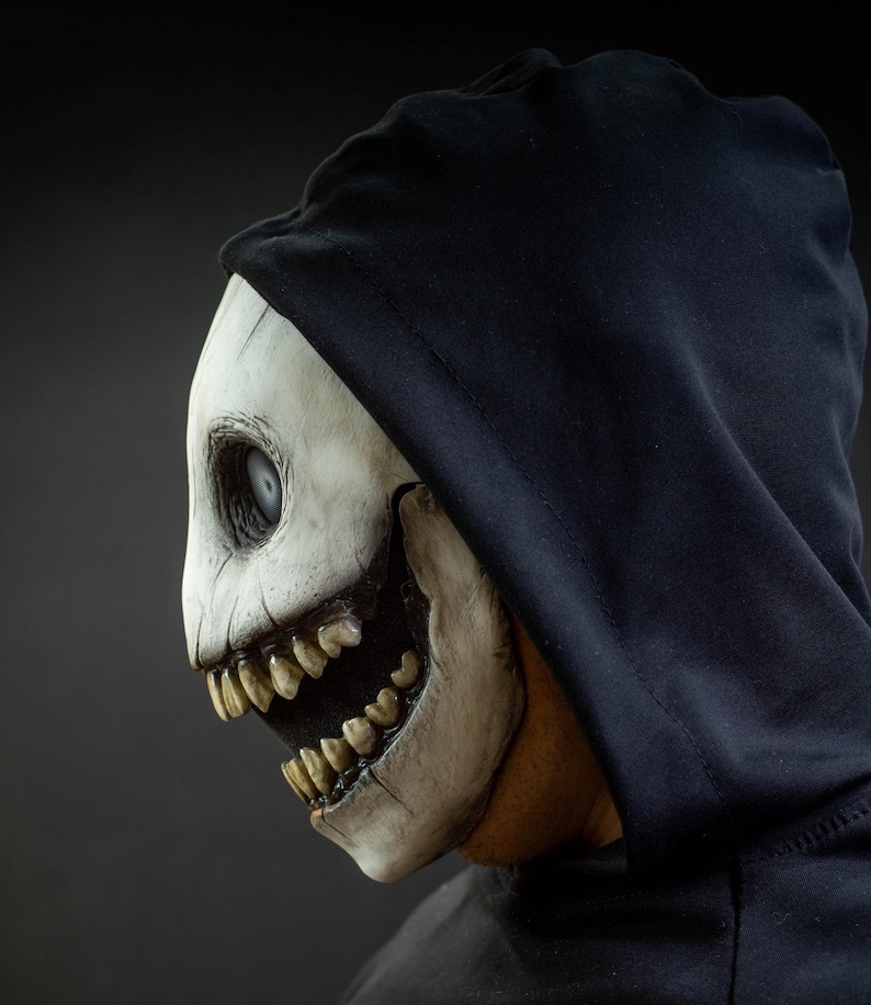 May include: A side profile of a white skull mask with a black hood. The mask has a wide open mouth with sharp teeth and a single, large eye. The mask is worn over a black hooded sweatshirt.