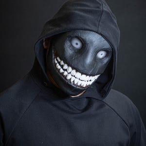 May include: A person wearing a black hoodie with a black mask that has a wide, toothy grin and white eyes.