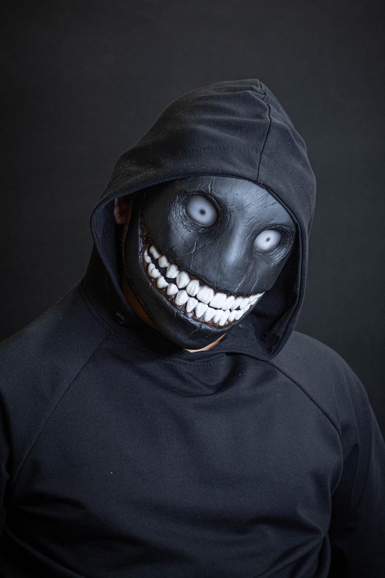 May include: A person wearing a black hoodie with a black mask that has a wide, toothy grin and white eyes.