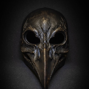 May include: A detailed, antique-style mask with a bird-like beak and large eye openings. The mask is dark bronze in color and has a textured surface, suggesting an aged or weathered appearance. The mask is likely for costume or decorative purposes.