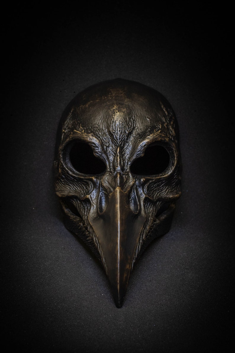 May include: A detailed, antique-style mask with a bird-like beak and large eye openings. The mask is dark bronze in color and has a textured surface, suggesting an aged or weathered appearance. The mask is likely for costume or decorative purposes.