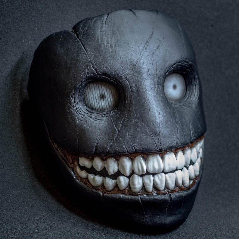 May include: A black creepy mask with a wide, toothy grin and white eyes. The mask is made of a textured material and has a realistic look.