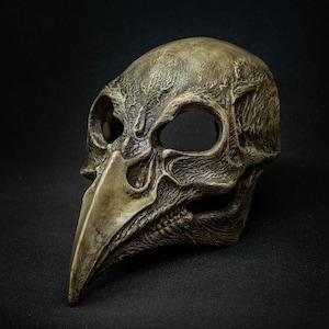 May include: A detailed, antique-style mask with a bird-like beak and skull-like features. The mask is primarily a weathered, muted brown with darker shading, suggesting age and texture. The eye sockets are large and empty, adding to the eerie aesthetic.