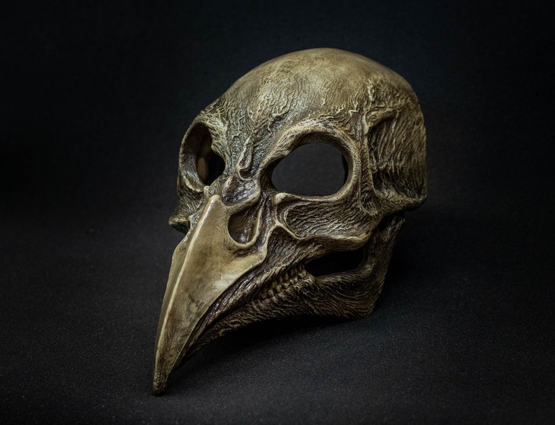 May include: A detailed, antique-style mask with a bird-like beak and skull-like features. The mask is primarily a weathered, muted brown with darker shading, suggesting age and texture. The eye sockets are large and empty, adding to the eerie aesthetic.