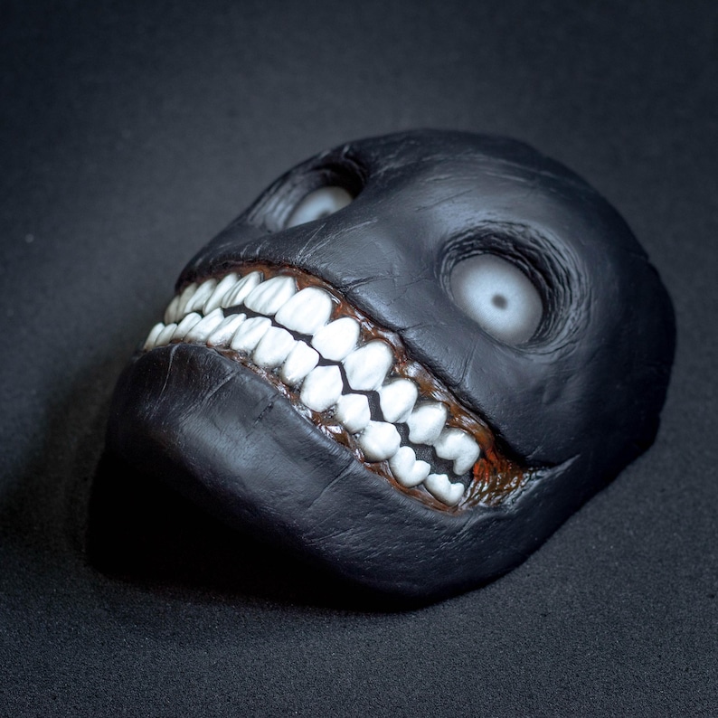 May include: A black leather mask with a wide, toothy grin and white eyes. The mask is designed to be scary and unsettling.