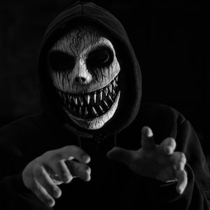 Op de afbeelding: Een zwarte hoodie gedragen met een wit en zwart masker met een gebarsten textuur, diepe holle oogkassen en een brede, tandige grijns met scherpe, puntige tanden. Het masker bedekt het gezicht en de capuchon is over het hoofd getrokken.