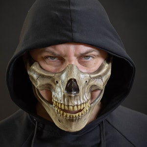Pu&ograve; includere: Una maschera a teschio beige con un design dettagliato, indossata con una felpa con cappuccio nera. La maschera copre la met&agrave; inferiore del viso, rivelando denti e lineamenti. Il tema generale &egrave; Halloween o costume.
