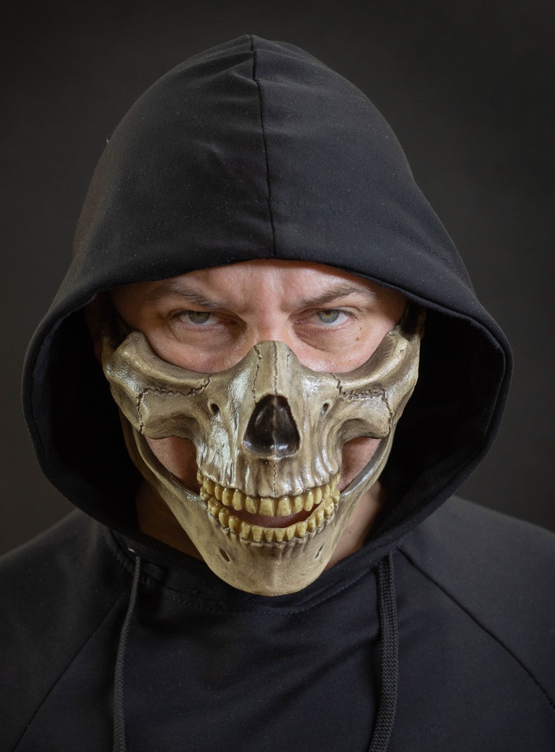 Pu&ograve; includere: Una maschera a teschio beige con un design dettagliato, indossata con una felpa con cappuccio nera. La maschera copre la met&agrave; inferiore del viso, rivelando denti e lineamenti. Il tema generale &egrave; Halloween o costume.