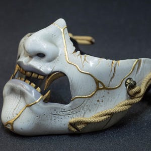 May include: A detailed, off-white Japanese Hannya mask with gold-colored accents. The mask features a demonic expression with visible teeth and a textured surface. A beige rope is attached to the side.