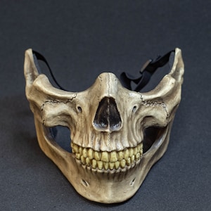 Pu&ograve; includere: Una maschera a mezza faccia a forma di teschio con un aspetto invecchiato simile all'osso. La maschera presenta una dettagliata raffigurazione di un teschio umano, compresi denti e cavit&agrave; nasale. Ha un cinturino nero regolabile.