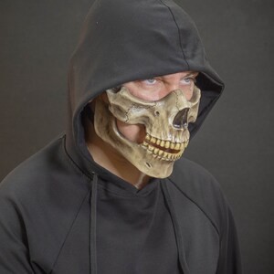 Pu&ograve; includere: Una maschera a teschio marrone e beige con una felpa con cappuccio nera. La maschera presenta dettagli, tra cui denti e orbite oculari. La felpa con cappuccio &egrave; nera e copre la testa.
