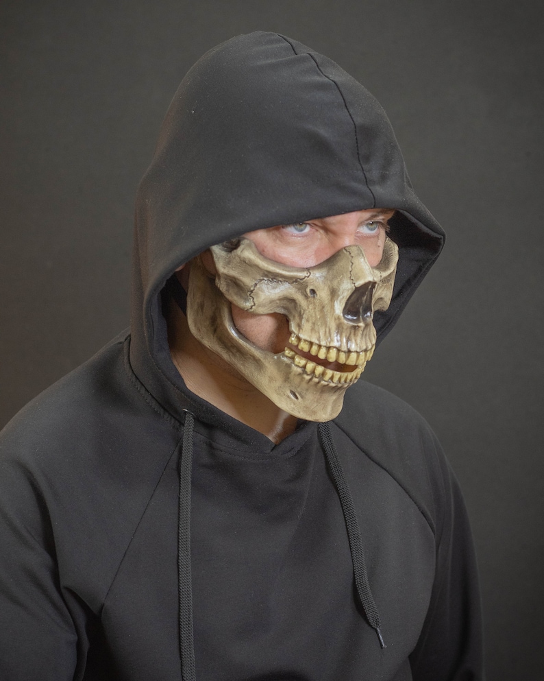 Pu&ograve; includere: Una maschera a teschio marrone e beige con una felpa con cappuccio nera. La maschera presenta dettagli, tra cui denti e orbite oculari. La felpa con cappuccio &egrave; nera e copre la testa.