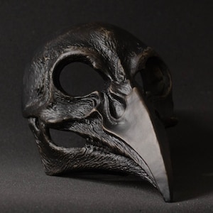 May include: A dark, textured mask with a bird-like beak and eye openings. The mask is a dark, matte color, possibly bronze or black, and has a detailed, weathered appearance, suggesting a gothic or historical theme.